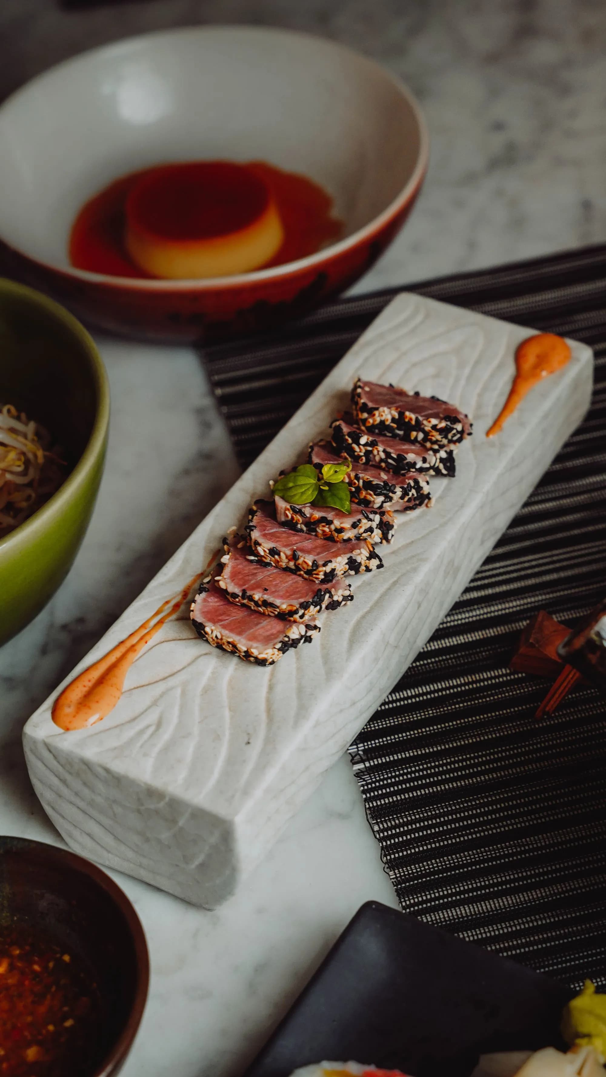 Lomo de atún rojo con costra de sésamo blanco y negro, sellado y cortado en láminas con el centro crudo y rojo, sobre una tabla de madera junto a un kamado cerámico encendido.