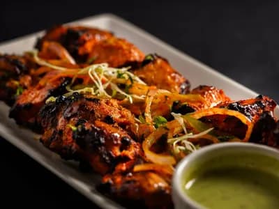 Contramuslos de pollo tandoori dorados y especiados sobre la parrilla de un kamado, junto a un cuenco de raita de pepino y gajos de lima.